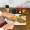 Person schneidet ein Brotlaib auf einem Glas Schneidebrett, umgeben von Tomate, Zwiebel, Knoblauch, Kräutern, Salzstreuer und einer Schale mit Öl.