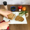 Person schneidet ein Brotlaib auf einem Glas Schneidebrett mit Marmoroptik, umgeben von frischem Gemüse, Kräutern und Küchenutensilien.