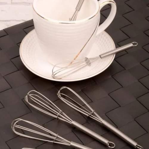 Sechs Edelstahl-Teelöffel in Schneebesenform, stilvoll auf einer schwarzen Oberfläche präsentiert, mit einer Teetasse im Hintergrund.
