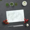 Glas Schneidebrett mit klassischem Marmor-Design auf einer dunklen Arbeitsfläche, umgeben von frischen Tomaten, Kräutern, Knoblauch, einem Messer und einem Küchentuch.