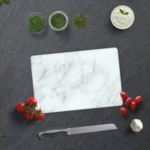 Glas Schneidebrett mit klassischem Marmor-Design auf einer dunklen Arbeitsfläche, umgeben von frischen Tomaten, Kräutern, Knoblauch, einem Messer und einem Küchentuch.