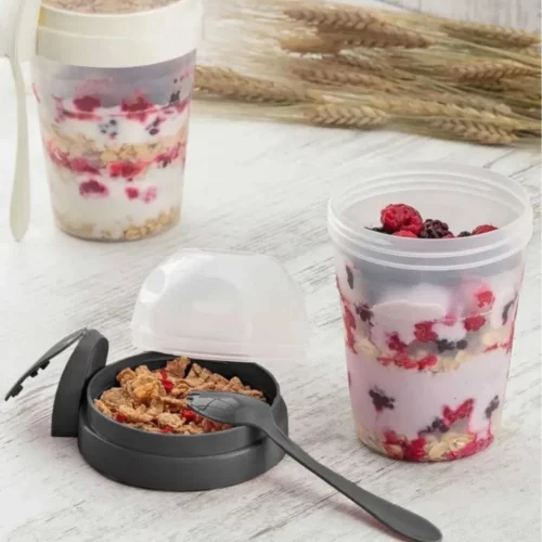 Zwei tragbare Joghurt- und Müslibecher mit Deckel und integriertem Löffel, gefüllt mit Joghurt, Beeren und Müsli, auf einem hellen Holztisch.