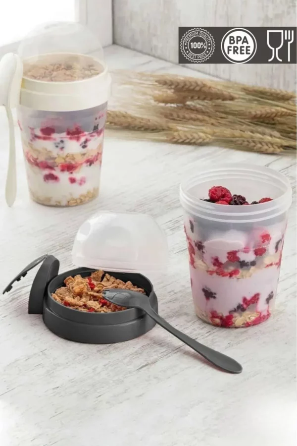 Zwei tragbare Joghurt- und Müslibecher mit Deckel und integriertem Löffel, gefüllt mit Joghurt, Beeren und Müsli, auf einem hellen Holztisch.